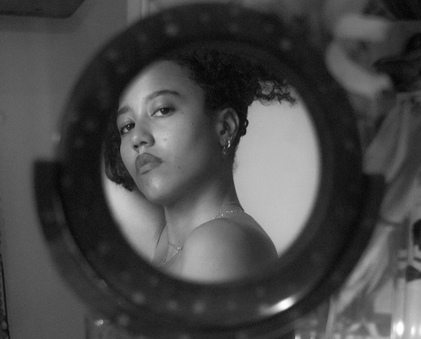 Black and white portrait of Care, a black trans* non-binary person, in a small circular mirror. Their shoulder is in view, and they are wearing an earring.
