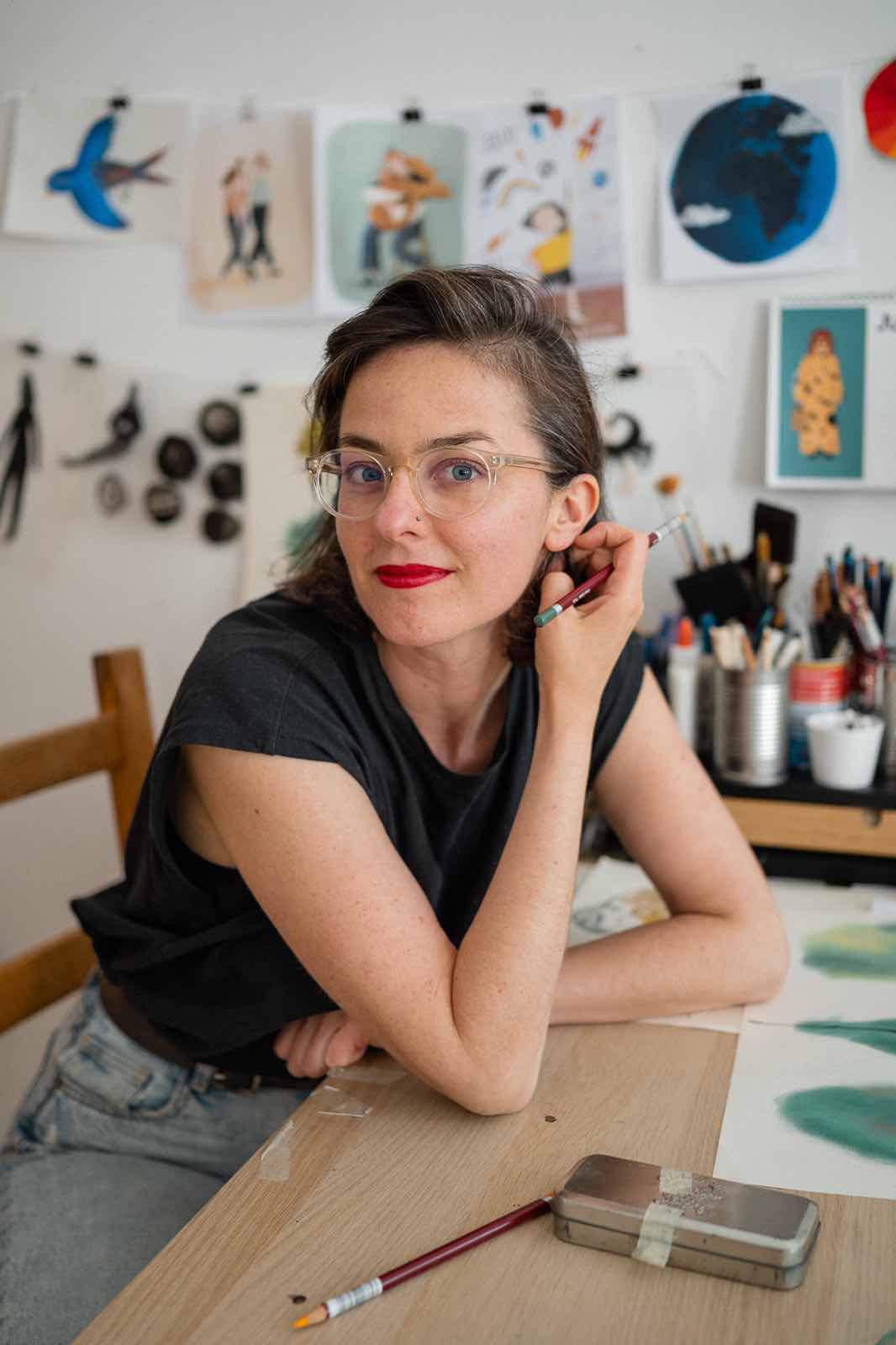 Portrait of Clara Gilod in her art studio. She is light skinned with dark brown hair, wearing clear glasses and bright red lipstick with a small smirk.
