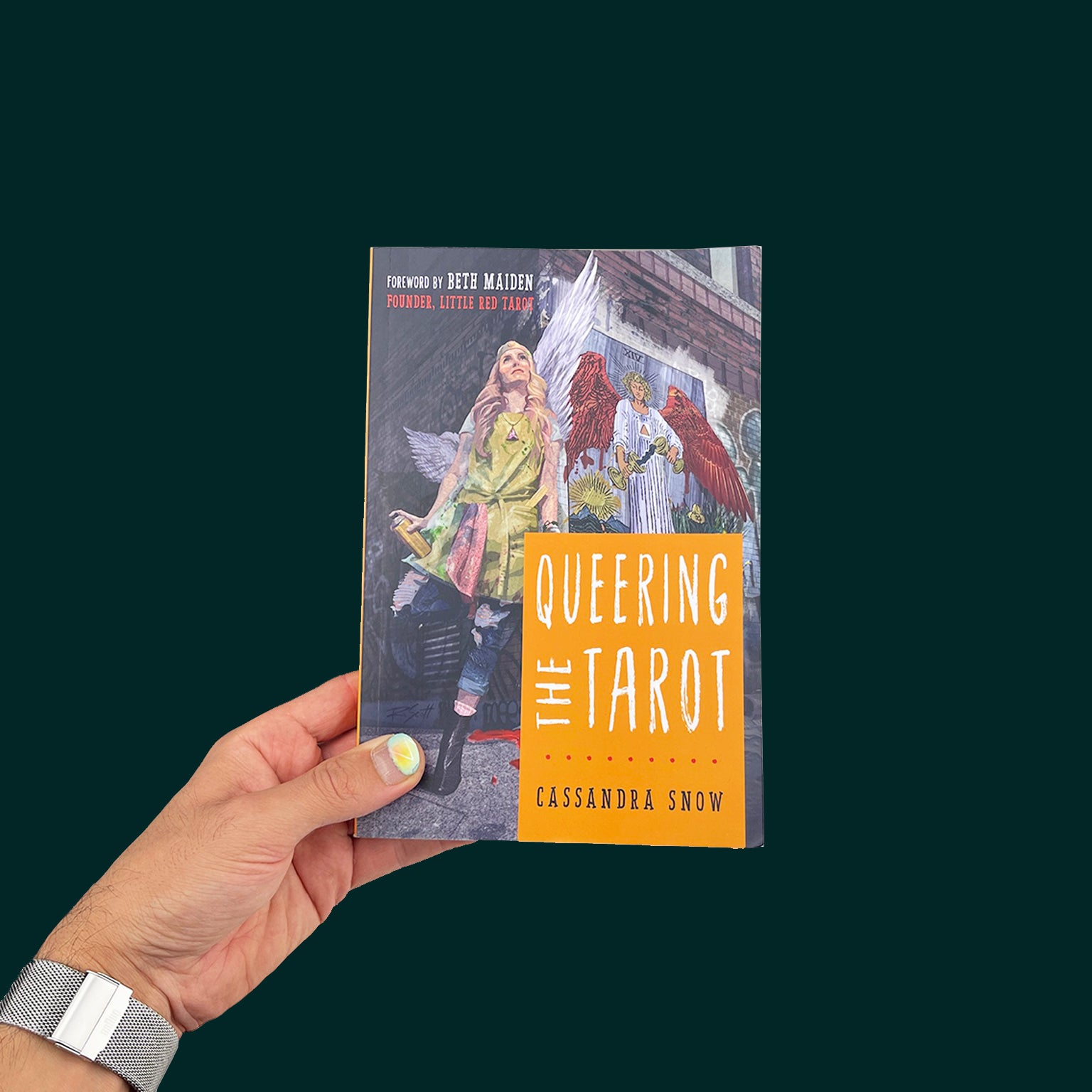 A hand with bright blue nail polish holds "Queering the Tarot" by Cassandra Snow. On the cover is an illustration of an angel walking away from a spray painted Temperance image on a brick wall, with a smile on her face.