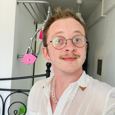 A selfie by the artist Rory Midhani. He's wearing a white collared shirt, thin black glasses, and has a moustache.