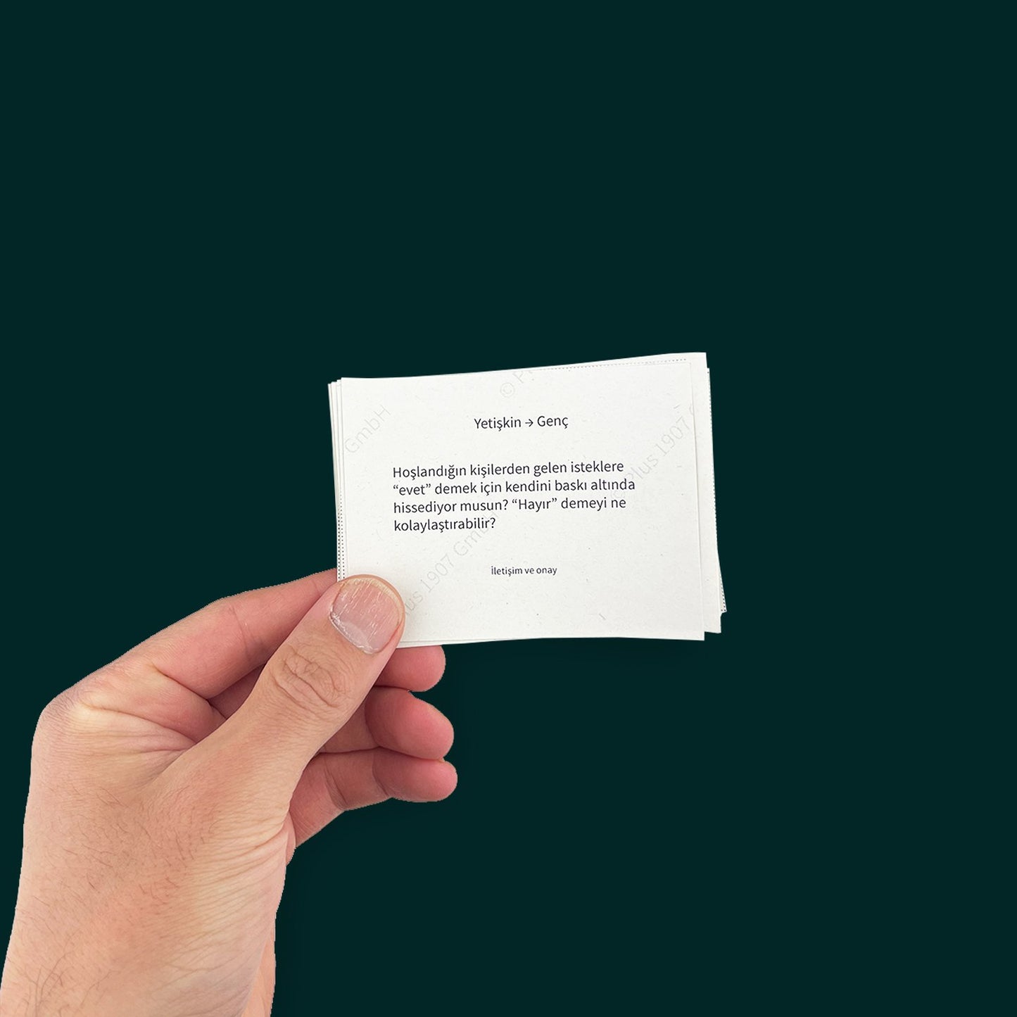 A left hand with holds a card with the text: "Yetiskin → Genç: Hoslandigin kisilerden gelen isteklere
"evet" demek için kendini baski altinda hissediyor musun? "Hayir" demeyi ne kolaylastirabilir?" with the tagline "lletisim ve onay" below. Against a deep green background.