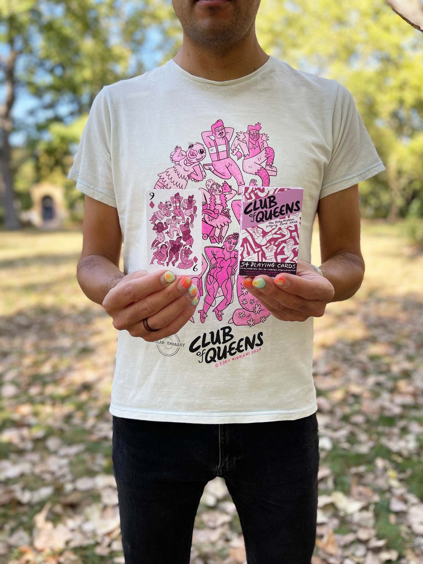 Person holding Club of Queens queer playing cards and wearing matching upcycled t-shirt with drag queen art