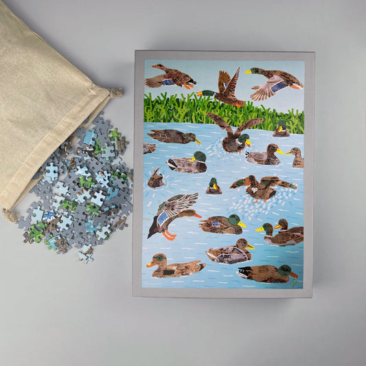 1000-piece jigsaw puzzle of trans*, inter*, non-binary ducks in pond, pieces spilling from cotton bag beside box.