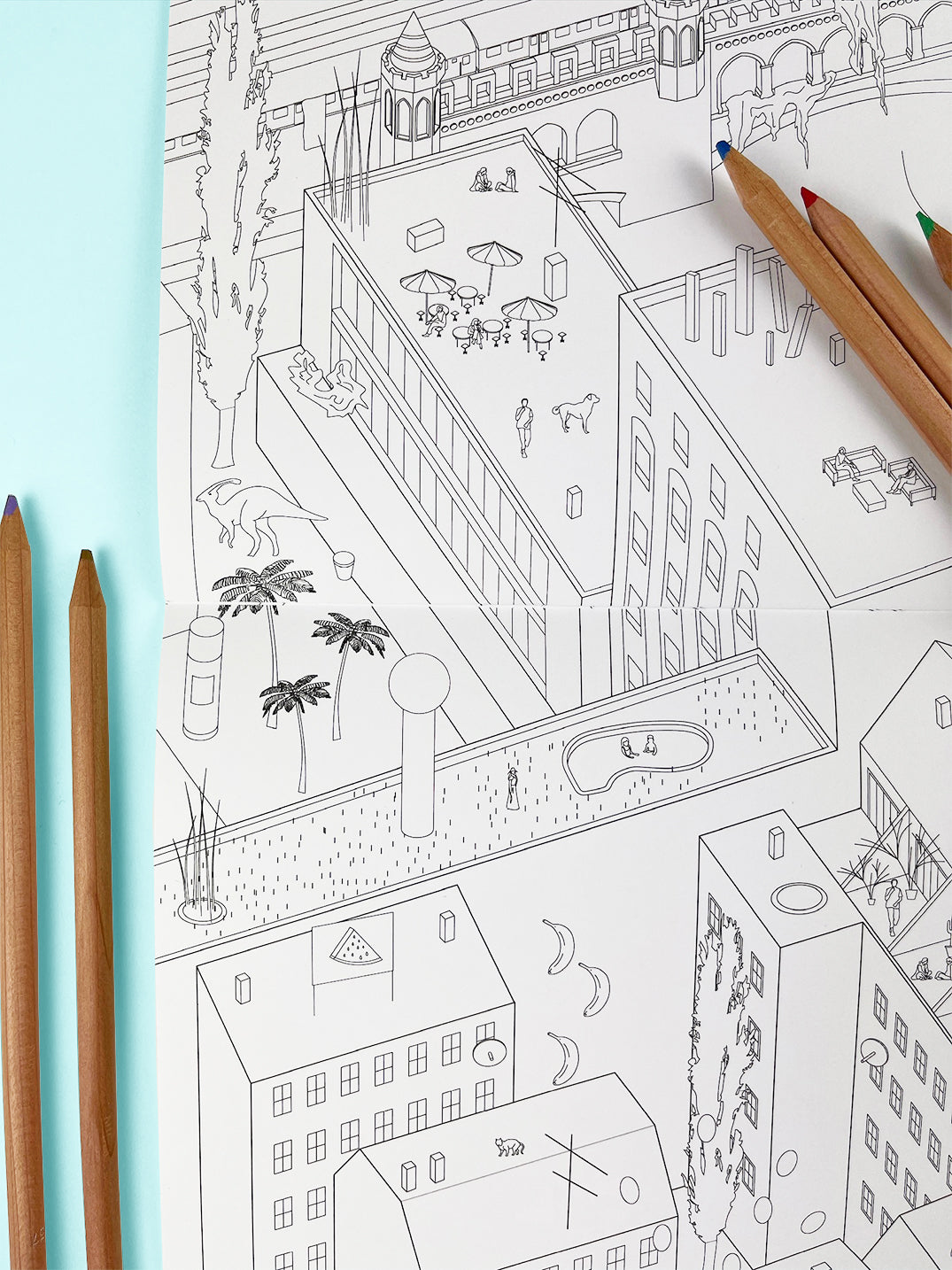 Photo of coloring book on a light blue background with colored pencils. The scene in the book is Kreuzberg with dinosaurs and whimsical elements.