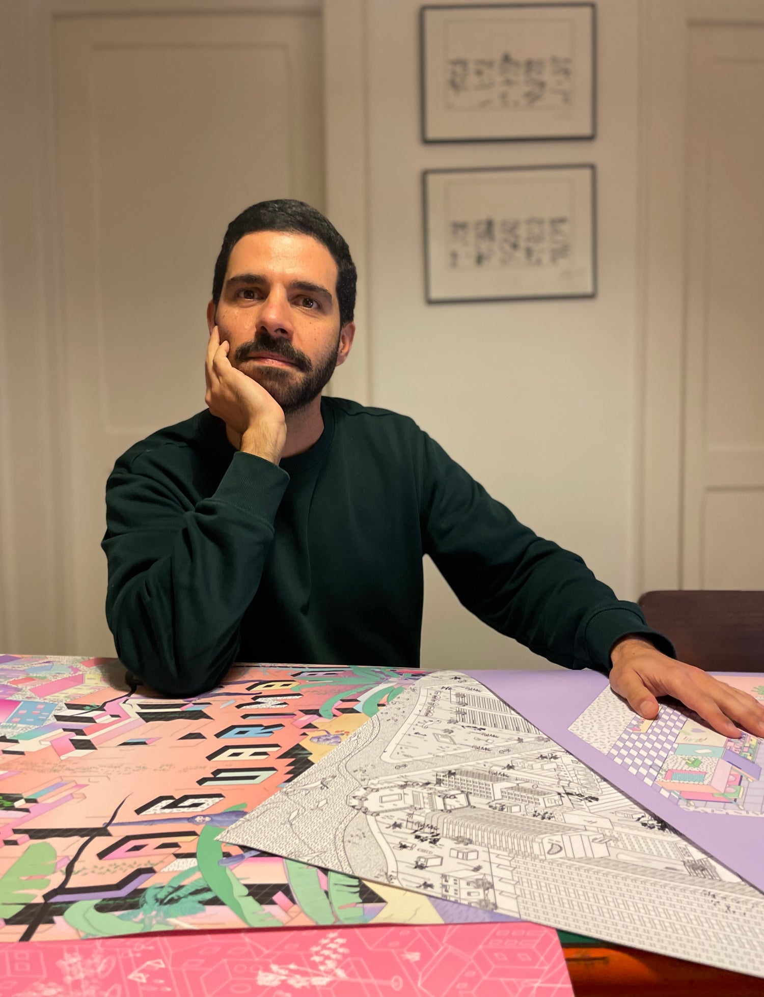 Artist and architect Bruno Pinto da Cruz sits at a table with his colourful, geometric designs on the table. He is wearing a black sweater, with one hand on the table, and the other holding his chin.