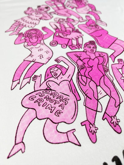 Detail photograph of bright pink illustration of drag queens on a white cotton shit. The drag queen closest, on the bottom left, is wearing a dress that says "drag is not a crime" and has her eyes closed.