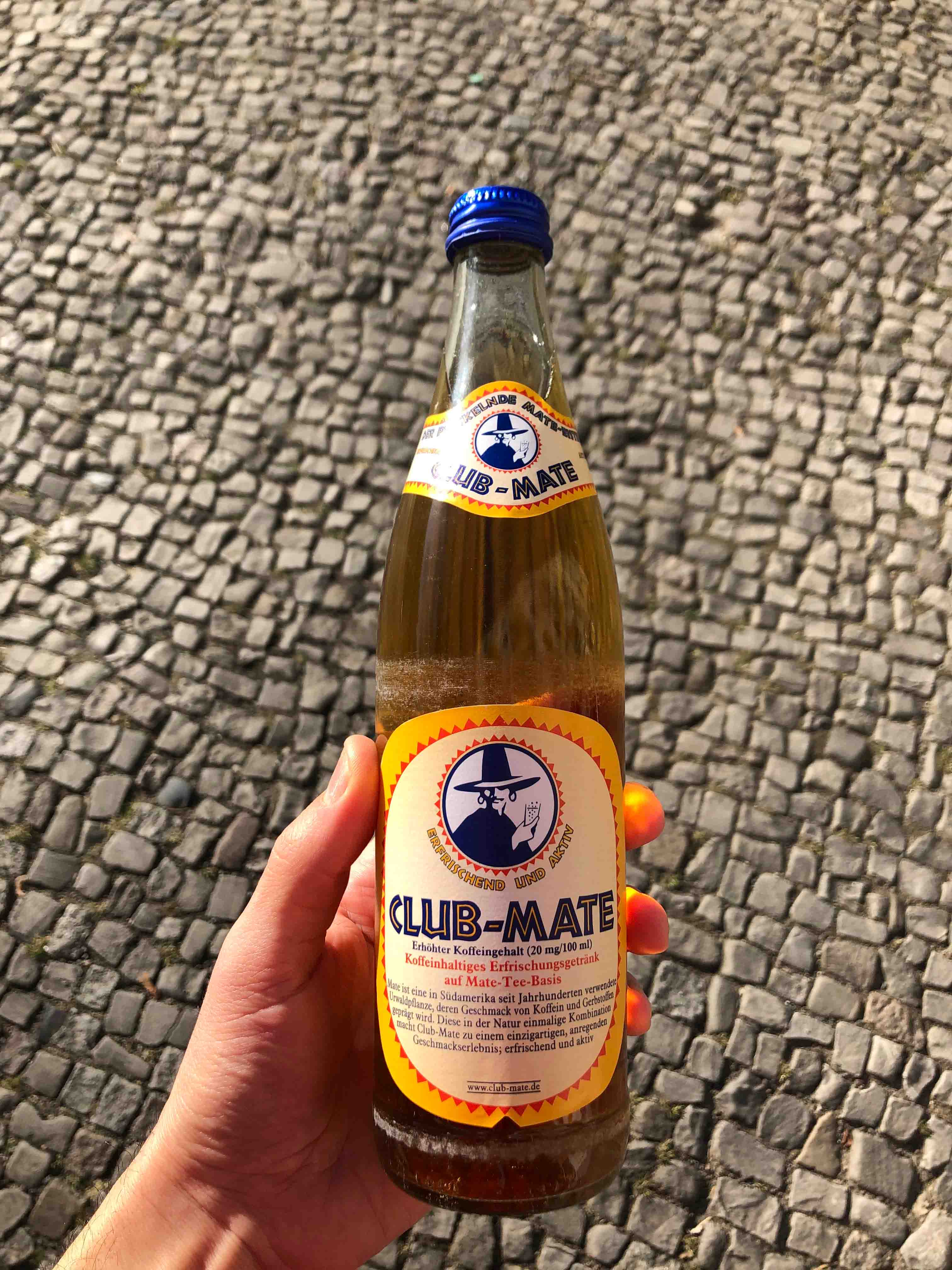 Hand holding a Club Mate bottle with a cobblestone sidewalk in the background.