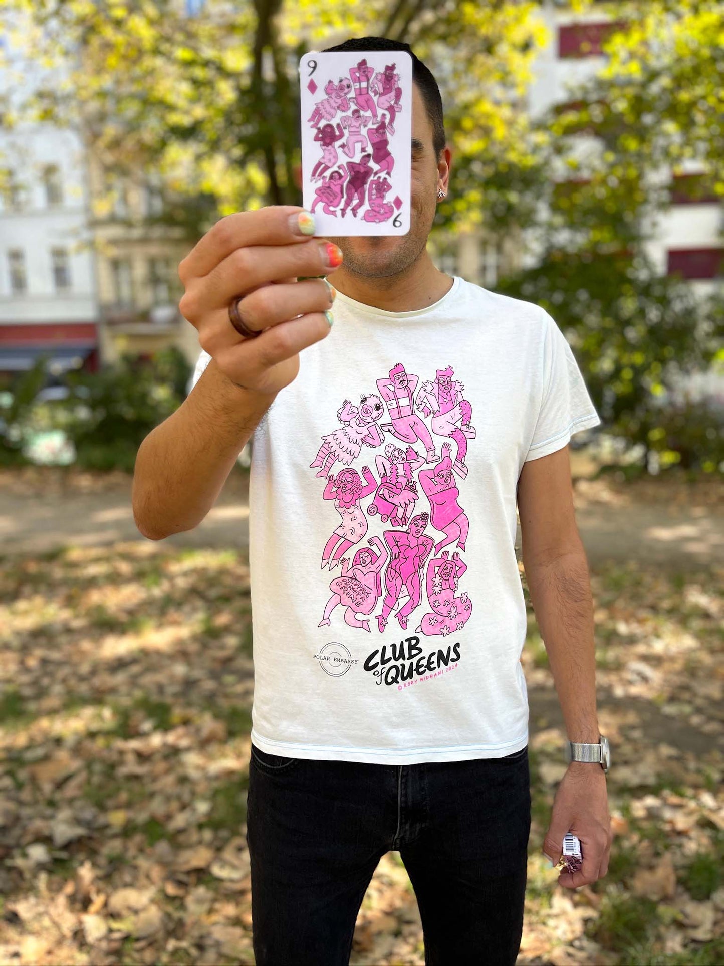 Person holding the 9 of Diamonds card from Rory Midhani's playing cards Club of Queens with a Graphic Tee with the drag queens from the playing card standing in a park in Berlin