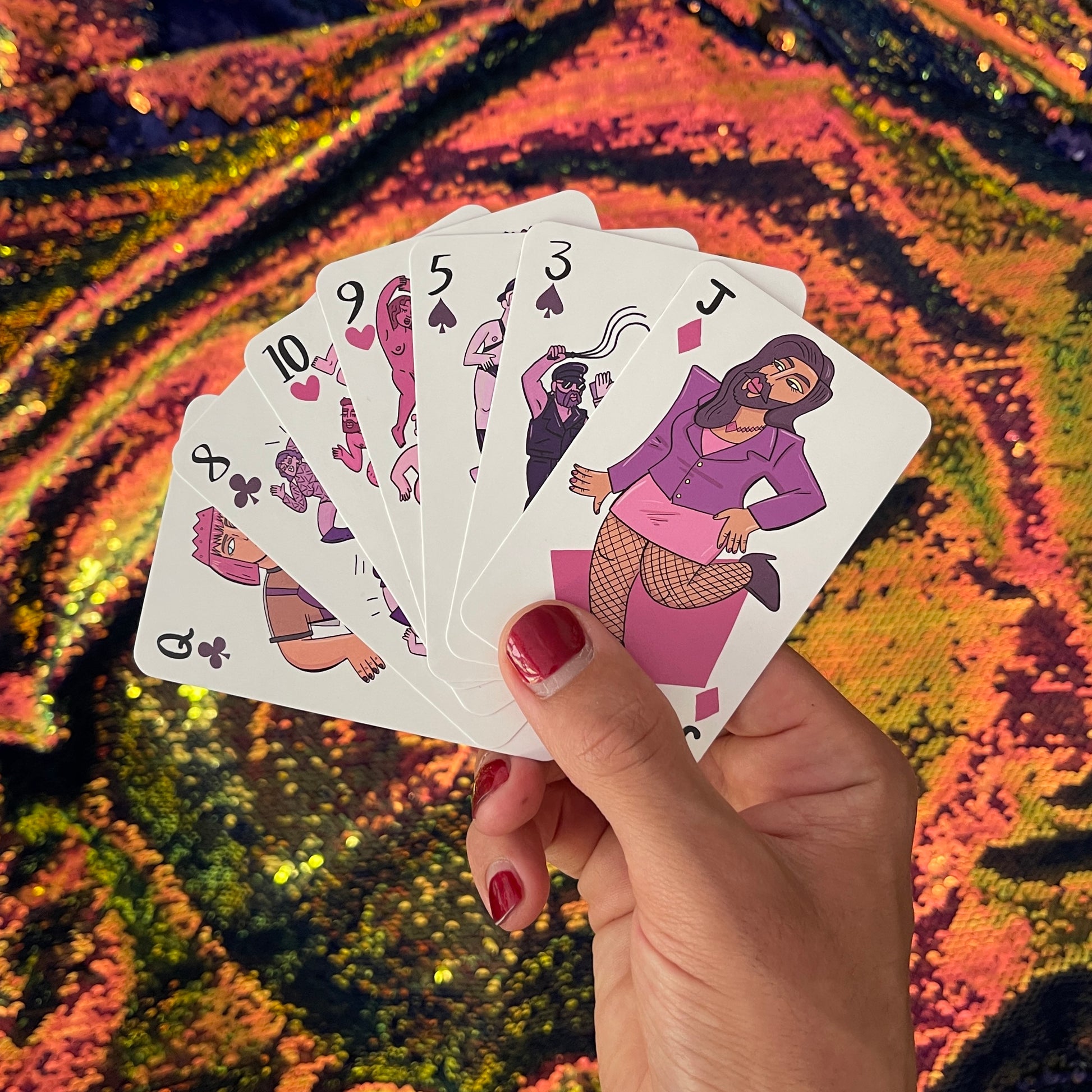 A hand with red nail polish holds up a hand from Rory Midhani's Club of Queens playing card deck, against a gorgeous sequin cloth background. The top card is a Joker, with an illustrated character in a blazer, skirt with leggings, heels, long hair, and a beard.