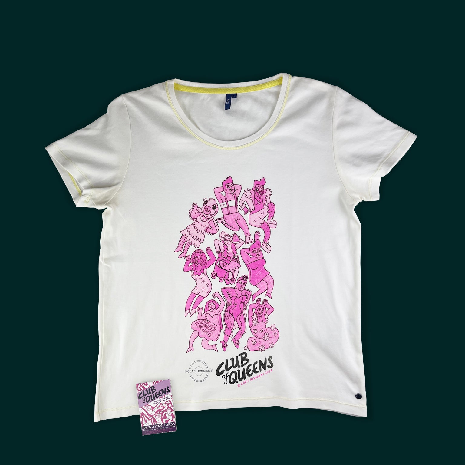 A deck of cards titled "Club of Queens" sits on a white shirt with a graphic from the deck of cards: nine bright pink drag king and queens, including one in a wheelchair, one with a bird costume, and one in a dress that says "Drag is not a crime".