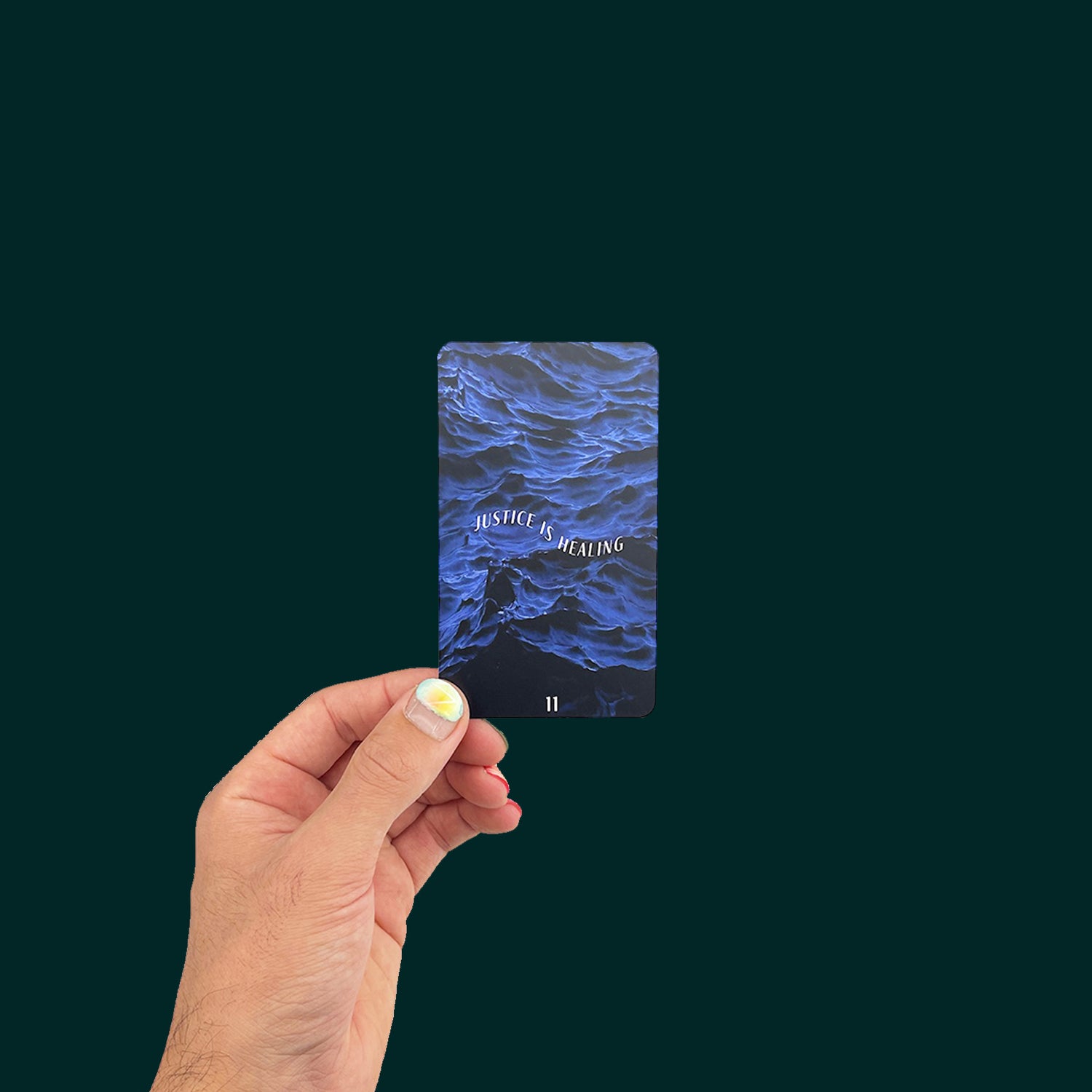 A hand holds a Tarot card from the Ancestral Healing Tarot deck, the Justice card, against a dark green background. The hand has bright pink and blue nail polish. On the card is dark blue image of waves, with the text "Justice is healing" in a wave form, with the number "11" at the bottom.