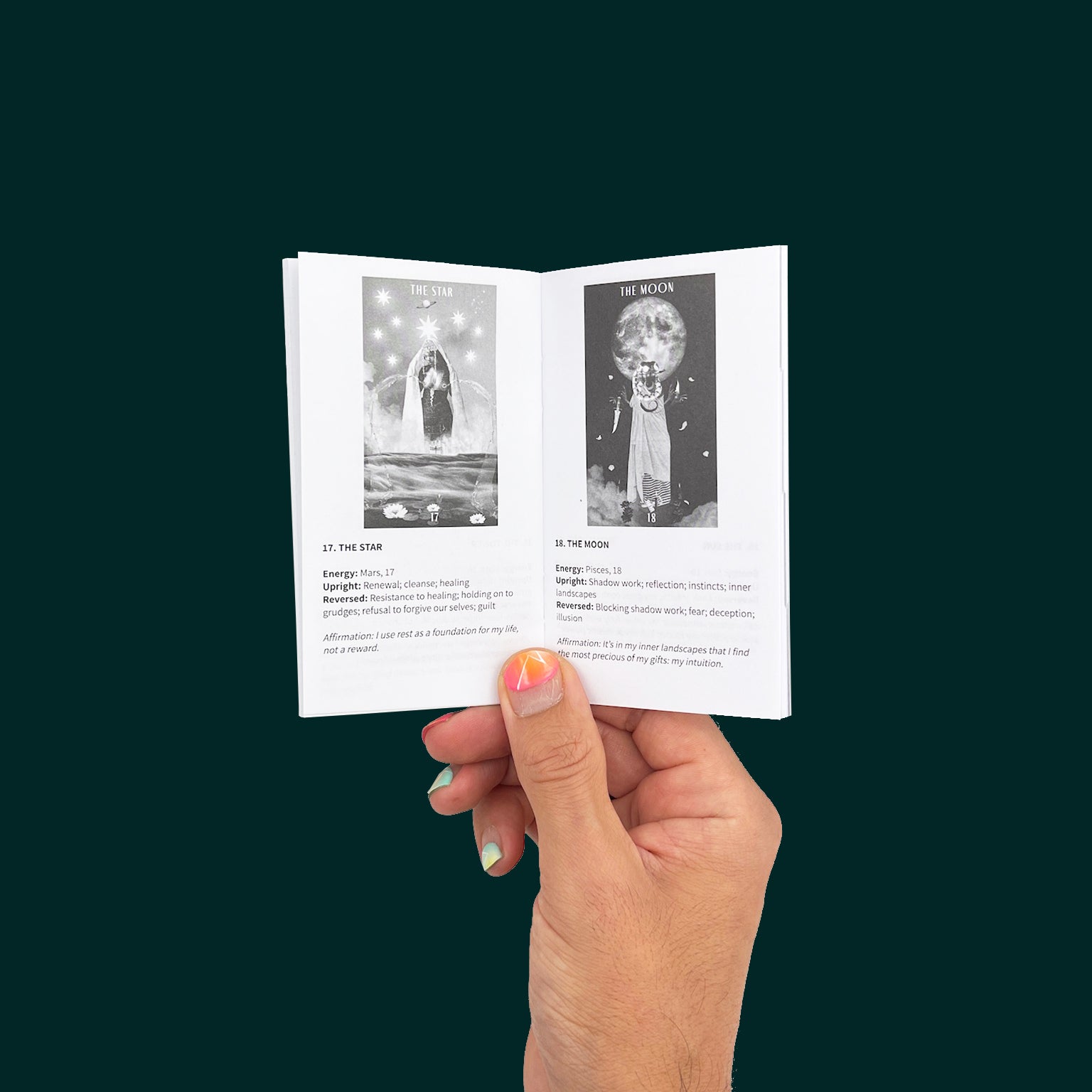 A hand with bright pink nail polish holds the guidebook from The Ancestral Healing Tarot Deck by Care for Polar Embassy. The black and white pages show The Star and The Moon cards.