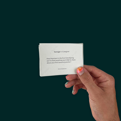 Hand holding print-at-home sex educaiton cards for families with teens. The hand has bright nail polish, and the question reads: "Teenager → Caregiver
How important is the first time having sex? Is there anything you'd like to share about your first sexual encounter?" with the tagline "Desire & pleasure" at the bottom. Against a dark green background.