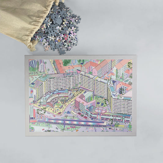 Puzzle of a colorful illustration of Kottbusser Tor, Berlin Kreuzberg, against a gray background. The puzzle pieces are coming out of a cotton bag on the table above the box.
