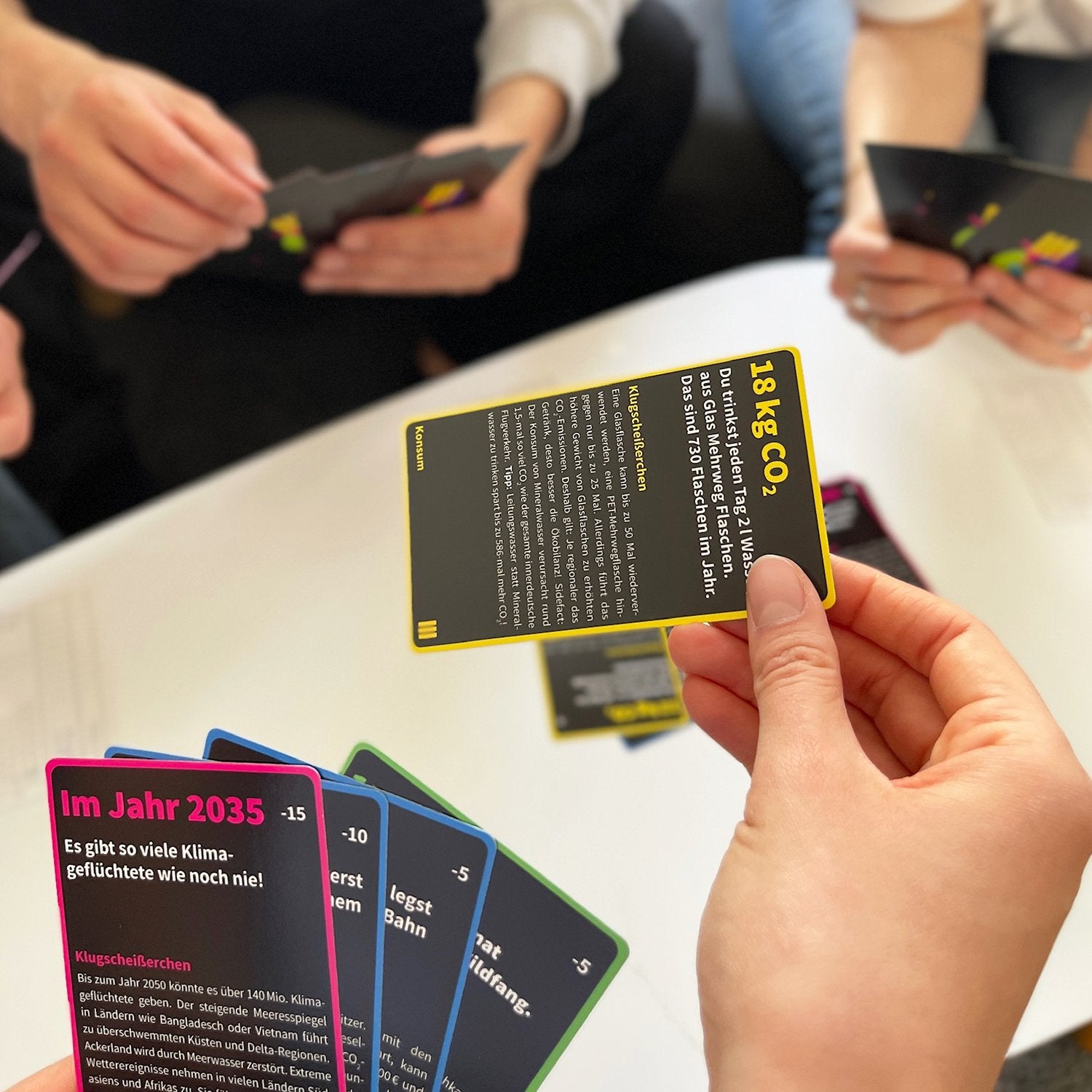 The card game Sinner or Saviour in play. A hand holds out a black card with yellow text and border, playing the card. Hands are holding playing cards in the background. 