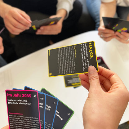 The card game Sinner or Saviour in play. A hand holds out a black card with yellow text and border, playing the card. Hands are holding playing cards in the background. 