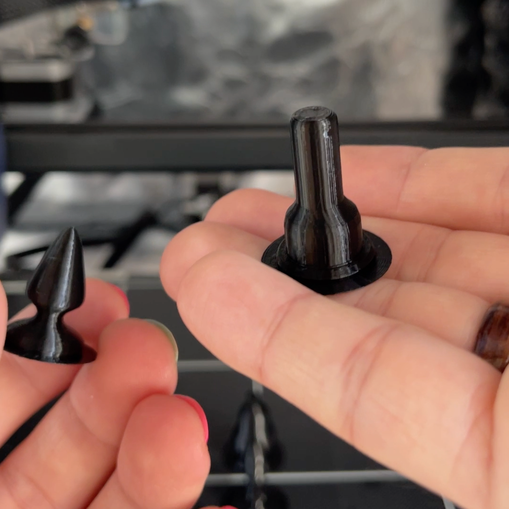 Hands holding black 3D printed chess pieces shaped like sex toys: a pawn and a rook.