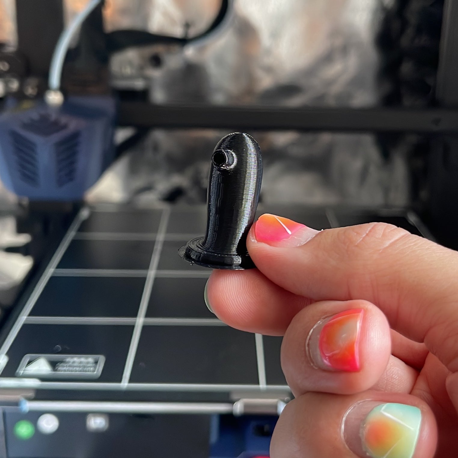 A hand with bright pink and green nail polish holding a black 3D printed chess piece (Knight) that looks like a suction sex toy.