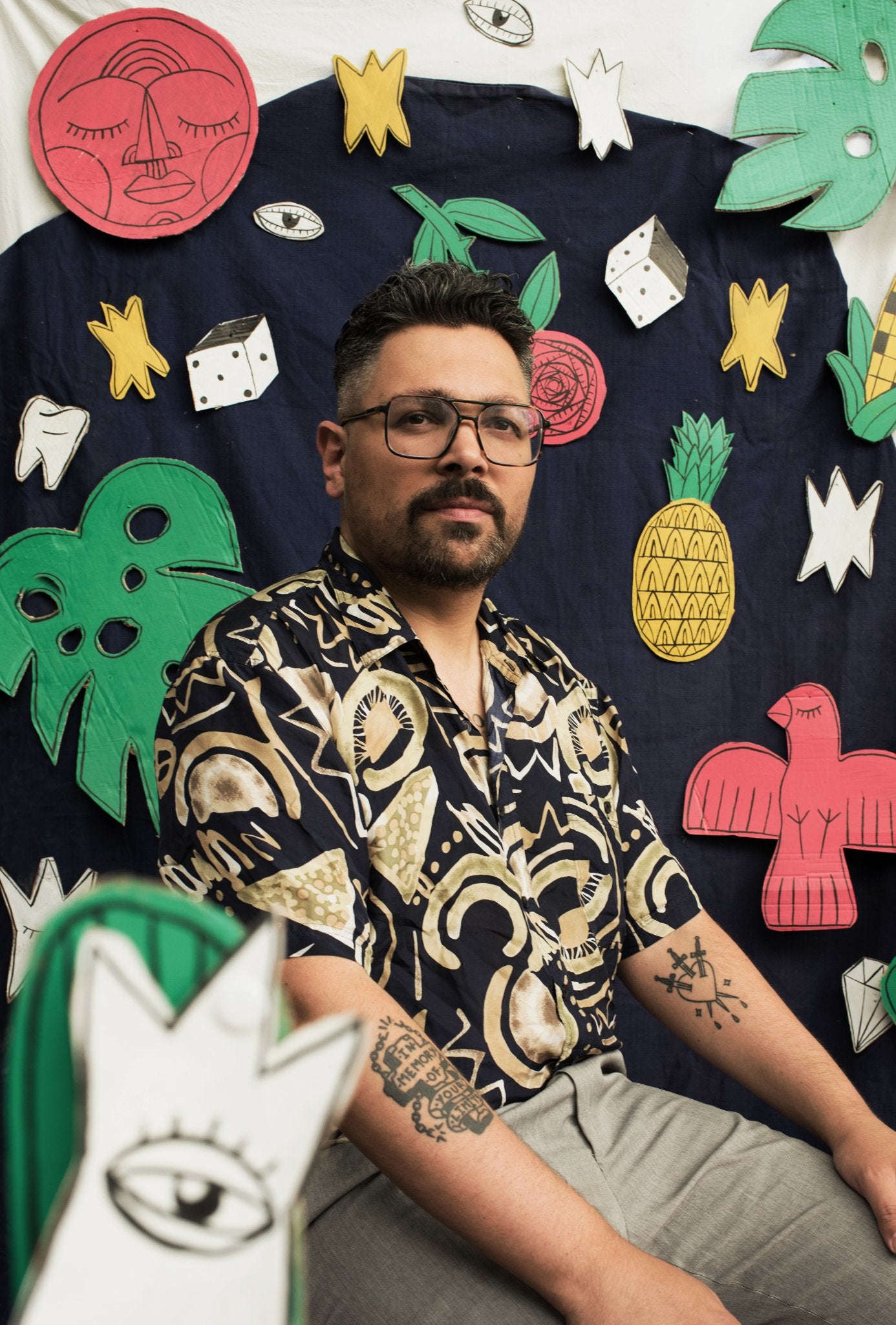 A portrait of the artist Denis Faneites. He sits wearing a flashy gold and black print shortsleeve shirt, and glasses. Behind him are colorful cutouts of his artwork, including pineapples, birds, dice, and stars. He gazes above the camera, and the overall effect makes him look playful, bold, and confident.