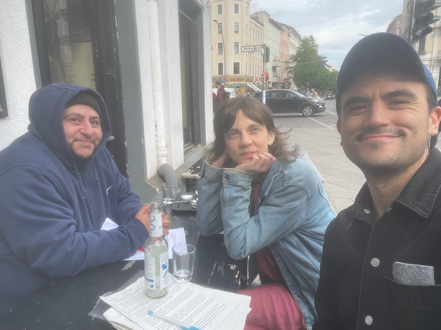 Three people sit at a table on a Berlin street. The person on the left is wearing a hoodie, and the two people on the right are wearing denim jackets.