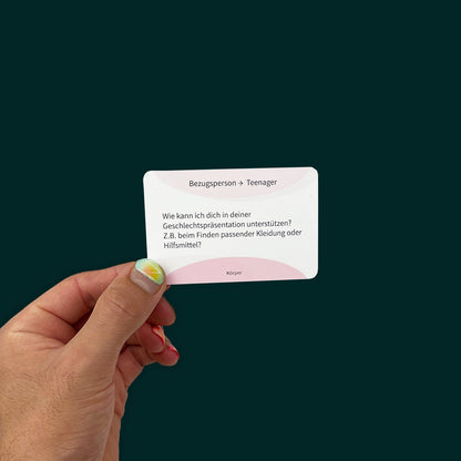 A hand with bright blue nail polish holds a card with the text: "Bezugsperson → Teenager: Wie kann ich dich in deiner Geschlechtspräsentation unterstützen? Z.B. beim Finden passender Kleidung oder Hilfsmittel?" with the tagline "Körper" below.
