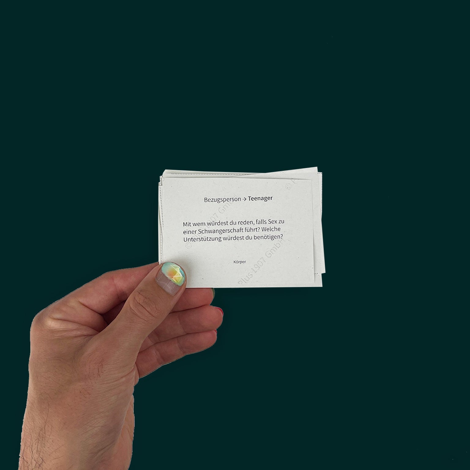 Hand holding print-at-home sex education cards for families with teens. The hand has bright nail polish, and the question reads: "Bezugsperson → Teenager
Mit wem würdest du reden, falls Sex zu einer Schwangerschaft führt? Welche Unterstützung würdest du benötigen?" with the tagline "Körper" at the bottom. Against a dark green background.