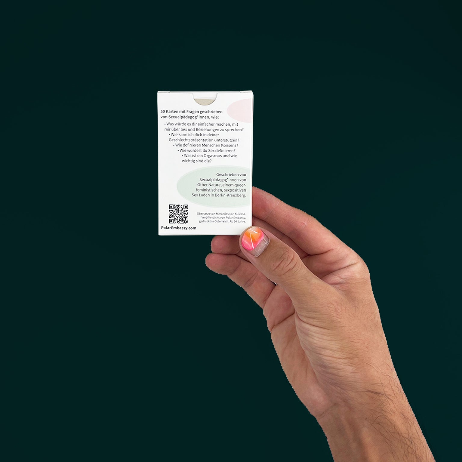 Hand holding a card deck of question cards for families with teens. The hand has bright nail polish, and the back of the deck says "50 Karten mit Fragen geschrieben von Sexualpädog*innen". Against a dark green background.