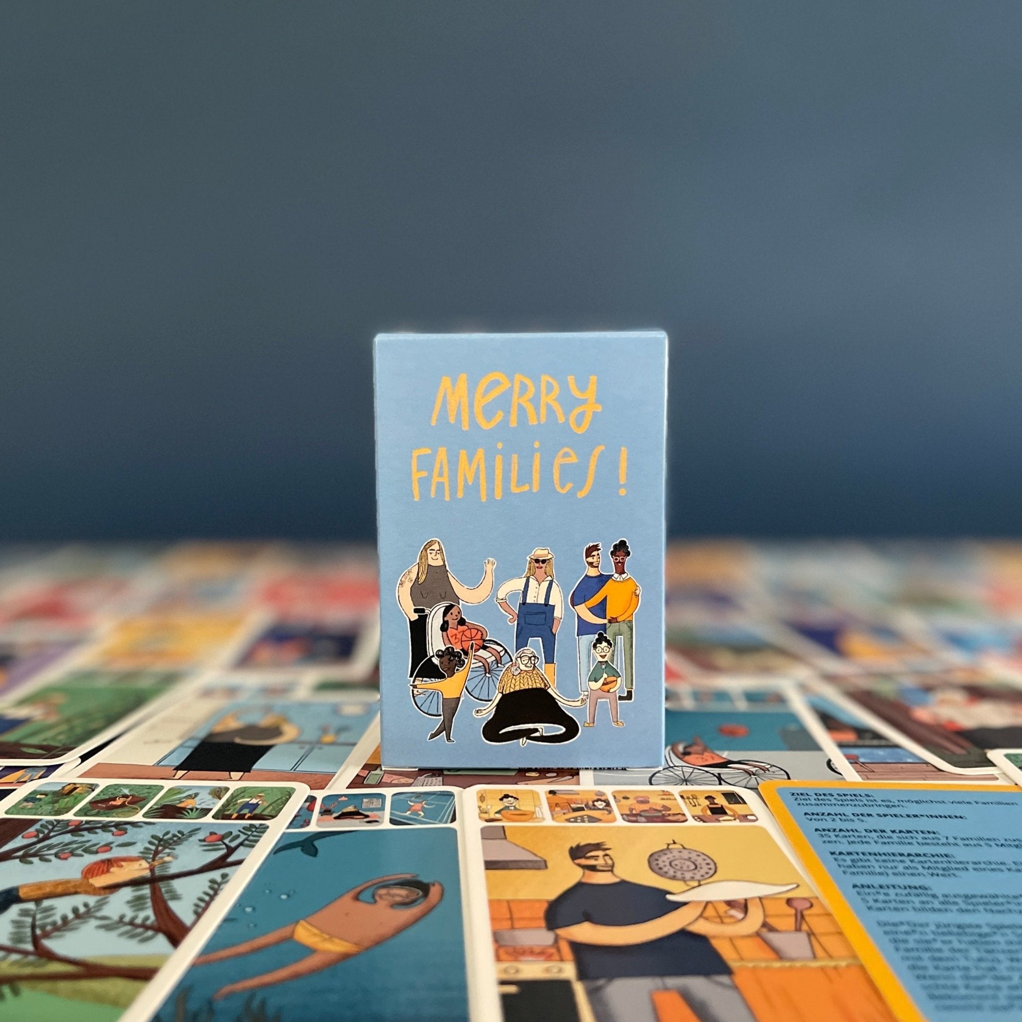Merry Families inclusive kids card game box standing on cards, against blue background.