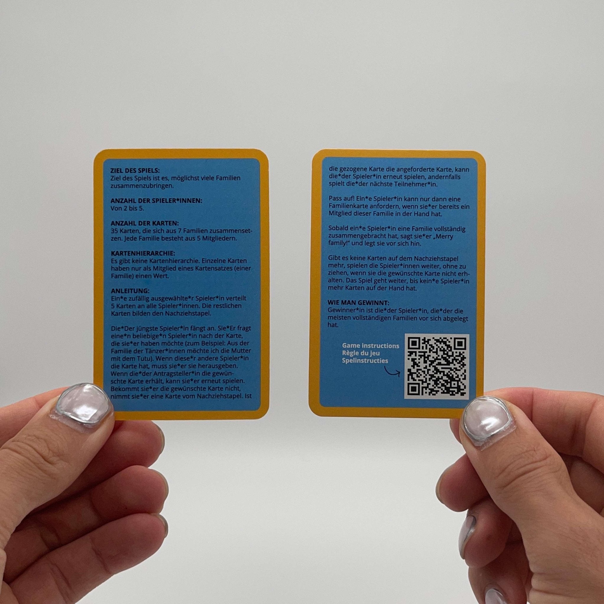 wo hands hold front and back of Merry Families kids card game instructions, silver and pearl nails.