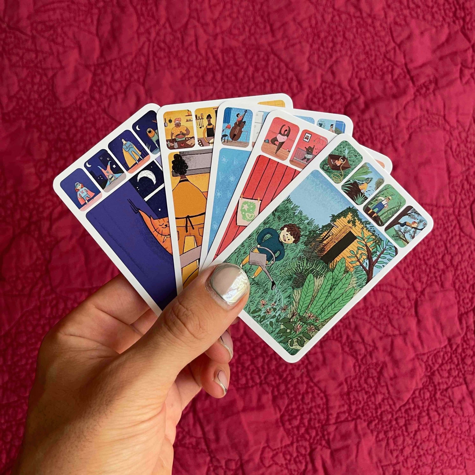A hand with gorgeous pearly and silver nail polish holds up five cards from the game Merry Families in their hand. The top card shows a small child watering the plants in a garden. Against a deep red cloth background.