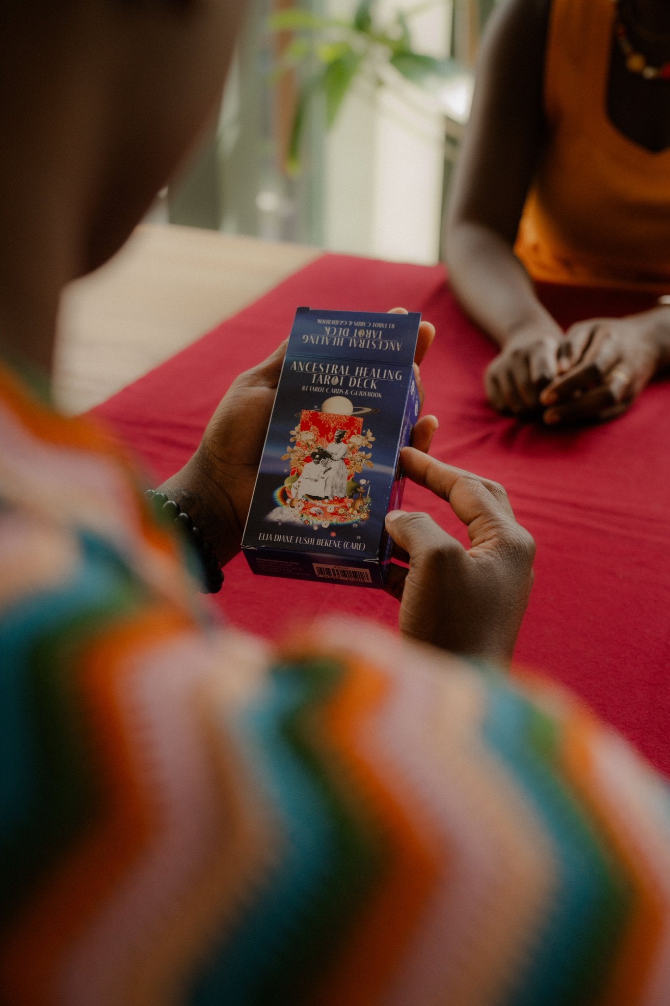 Hands holding the Ancestral Healing Tarot deck box, a collage art tarot deck by artist Care, over a deep-red cloth on a table.