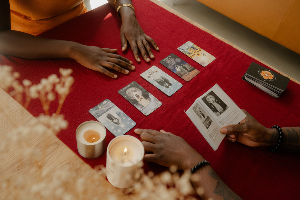 Two people share an Ancestral Healing Tarot reading at a candlelit table, cards spread on red cloth.