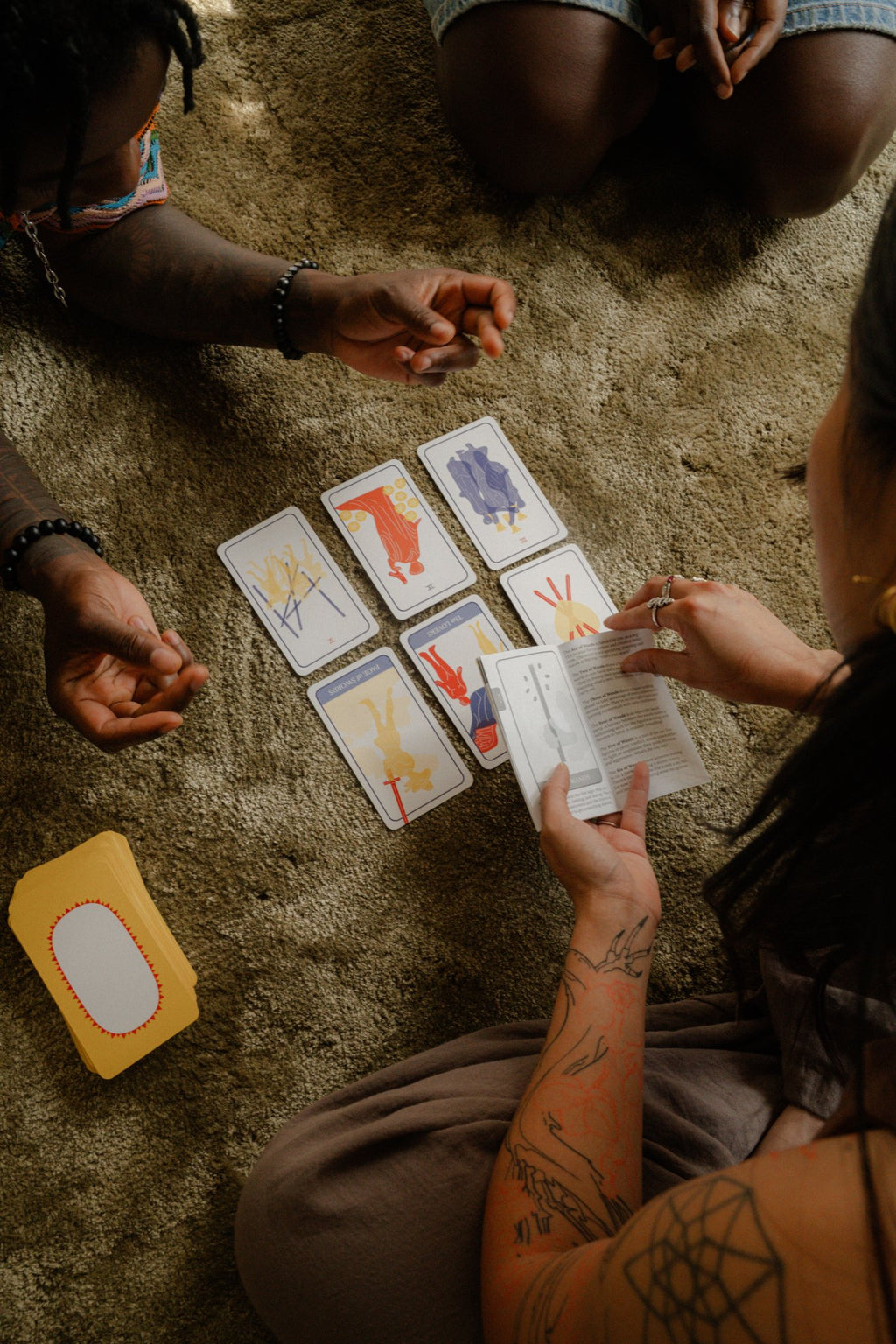 A tattooed reader interprets a 6-card spread from Polar Embassy's Club Tarot deck, guidebook open, as two people look on.