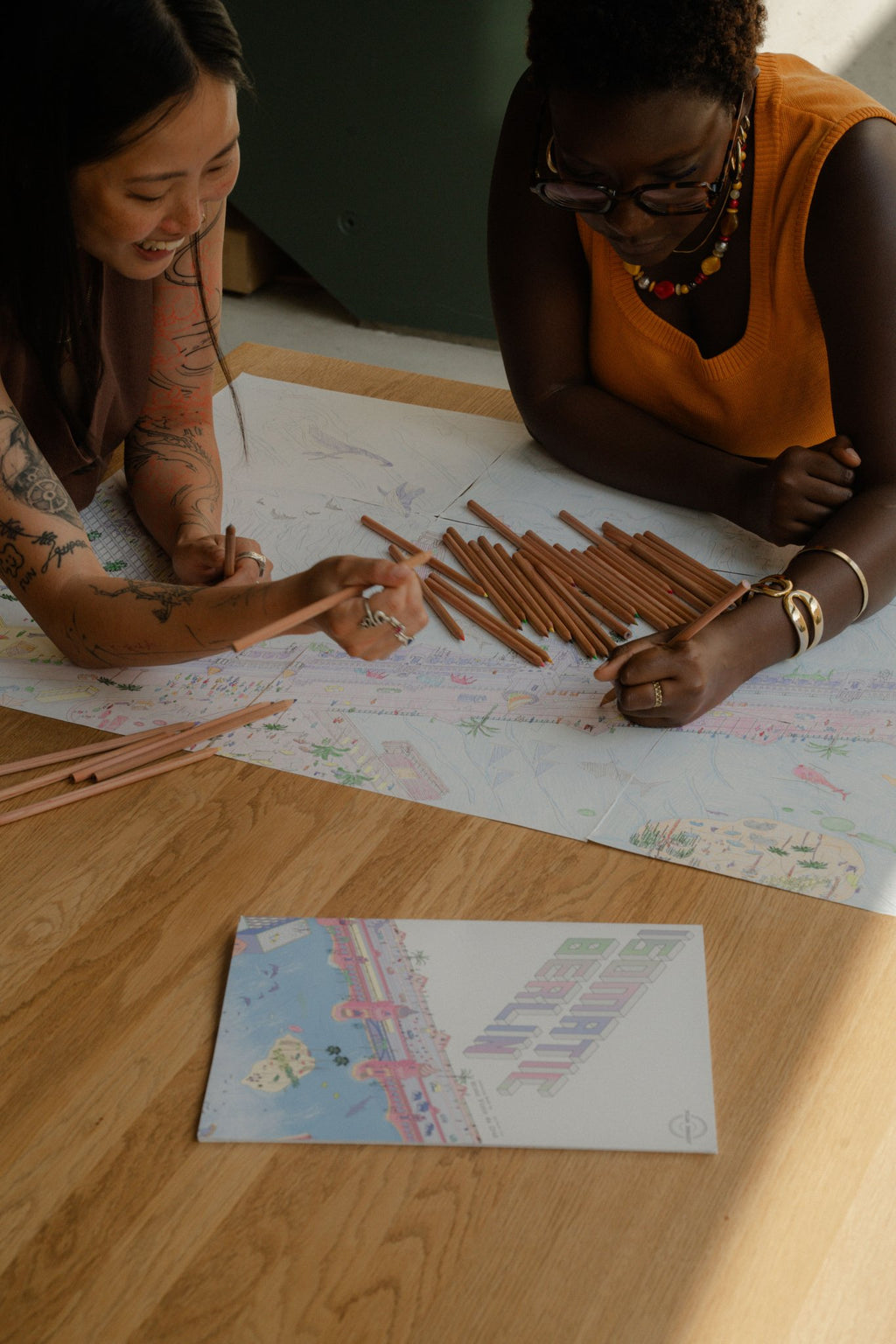 Two people colour the Berlin Coloring Book for adults at a wooden table, Isomatic art by Bruno Pinto da Cruz.