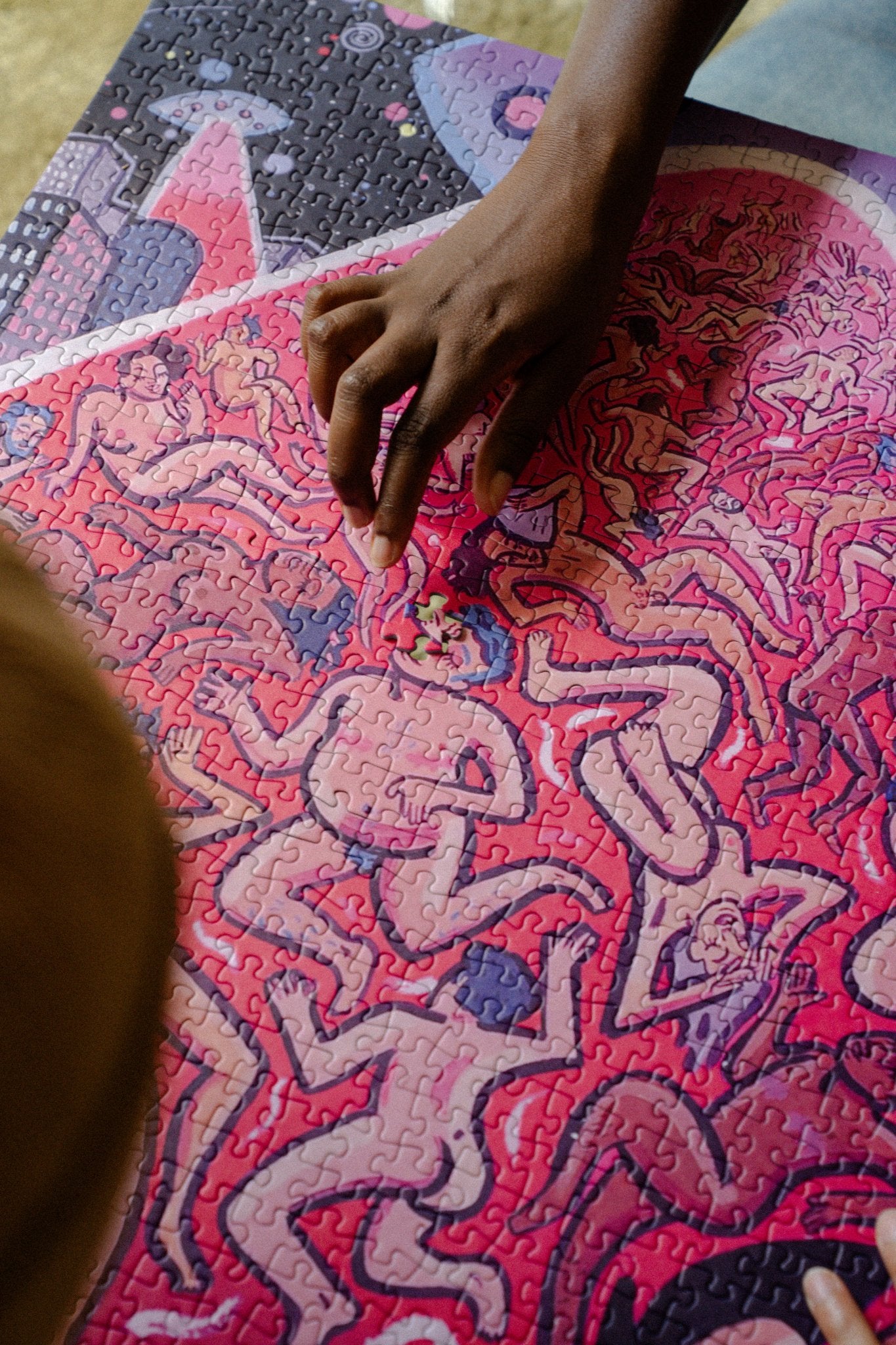 Hand placing the last piece into Rory Midhani's LGBTQIAUFO puzzle of nude trans and queer figures.