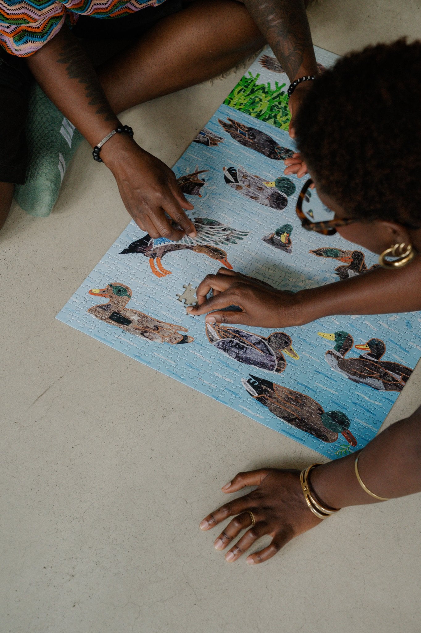 Two people finish TIN (trans inter non-binary) Ducks jigsaw puzzle, on a gray concrete floor.