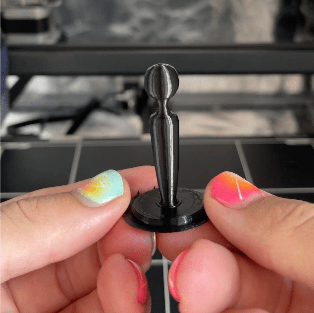 Hands holding a black 3D printed chess piece (king) shaped like a sex toy.