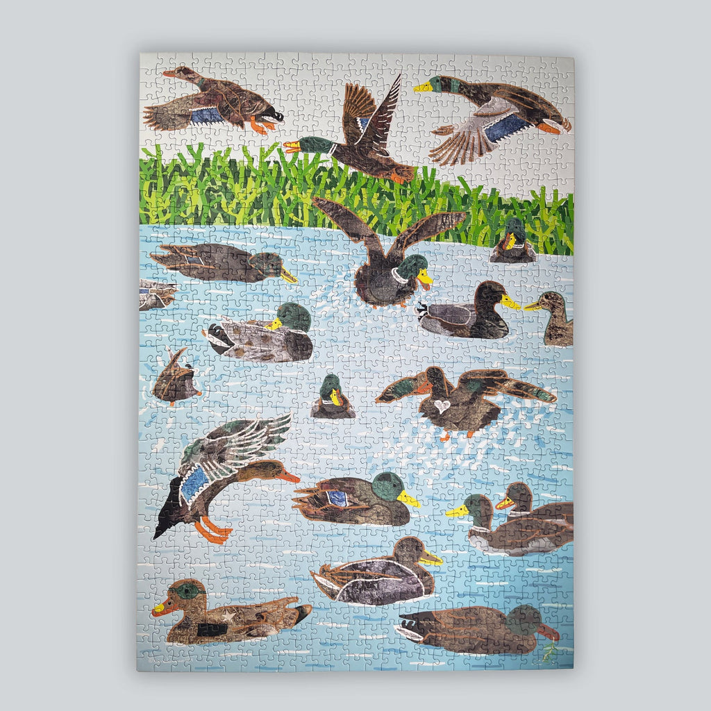 A rectangular jigsaw puzzle featuring a colorful duck scene, with trans*, inter, and non-binary mallards swimming and flying in blue water beneath green grass, shown against a light gray background.