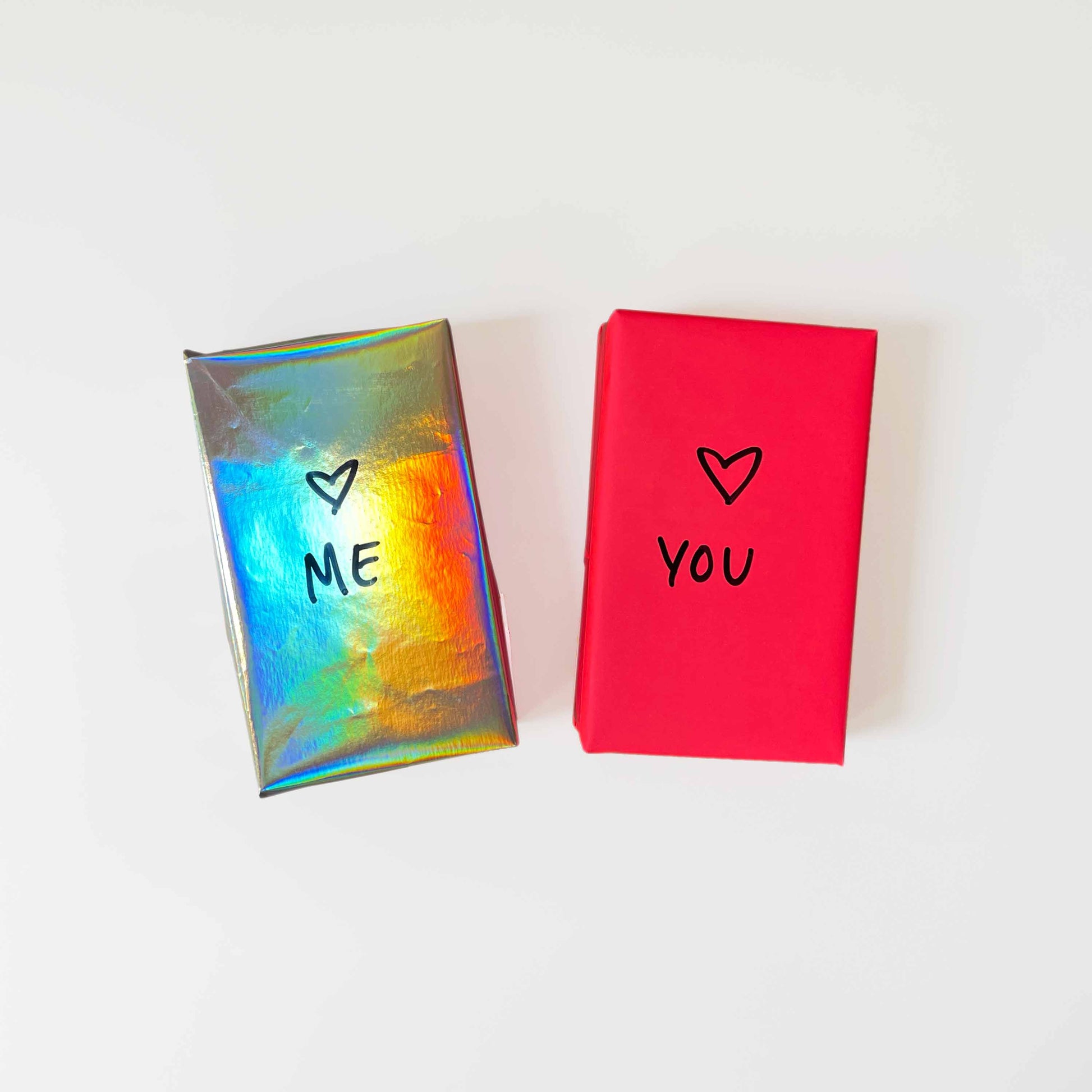 Two gift-wrapped decks of Tarot cards sit on a white background. The left one is in shiny rainbow paper with "ME" and a heart, and the one on the right is in deep pink wrapping paper with "YOU" and a heart.