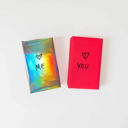 Two gift-wrapped decks of Tarot cards sit on a white background. The left one is in shiny rainbow paper with "ME" and a heart, and the one on the right is in deep pink wrapping paper with "YOU" and a heart.